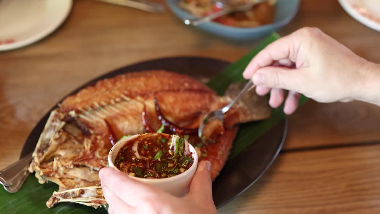 Hand pours spicy herb sauce over fried fish on banana leaf, natural daylight, close-up view