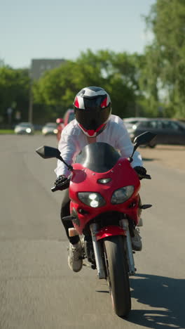 un motociclista con casco monta una bicicleta eléctrica roja rápidamente cerca de un camino de paseo, mientras que un coche rojo se mueve a lo largo, otros vehículos son visibles en la distancia con un poste eléctrico al lado