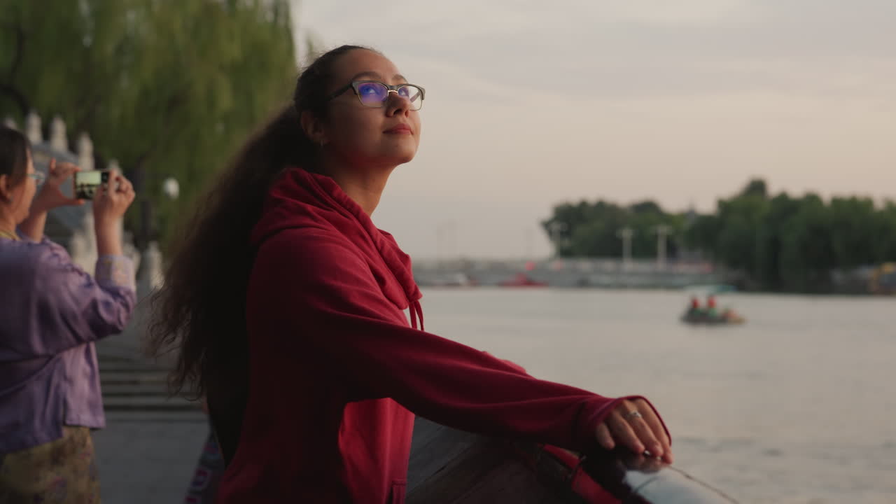 Young person gazes across river at sunset, side profile with glasses and red jacket, background tourists photographing boats and willow trees, calm water ripples and distant skyline, hopeful