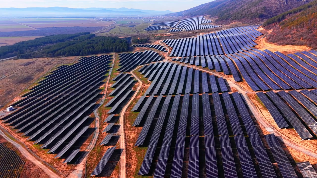 Stor solcellepark med dal og by i baggrunden, Bulgarien. Bredt droneperspektiv af solcellefelter i flere sektioner, rød jord, vinmarksrækker og en fjern bjergkæde.