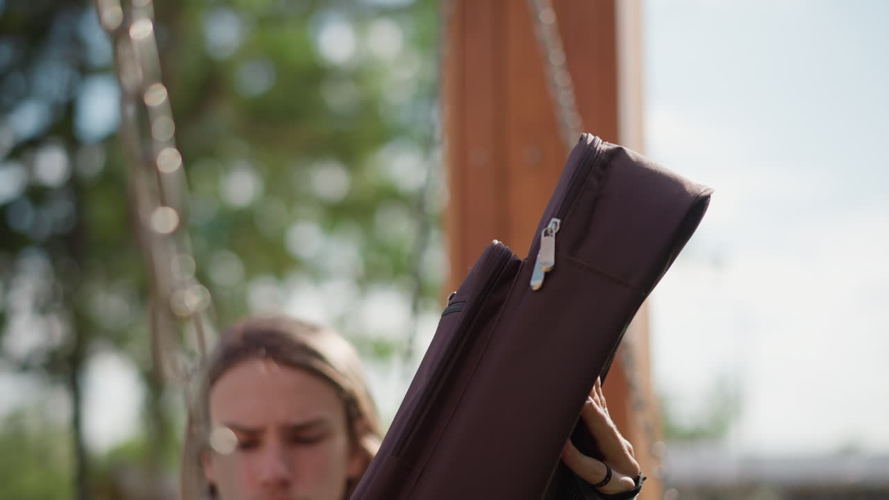 playground guitarist white man unzipping case, revealing acoustic guitar while adjusting strap and tuning, focused expression, denim jacket, chains and swing in background, warm daylight, candid