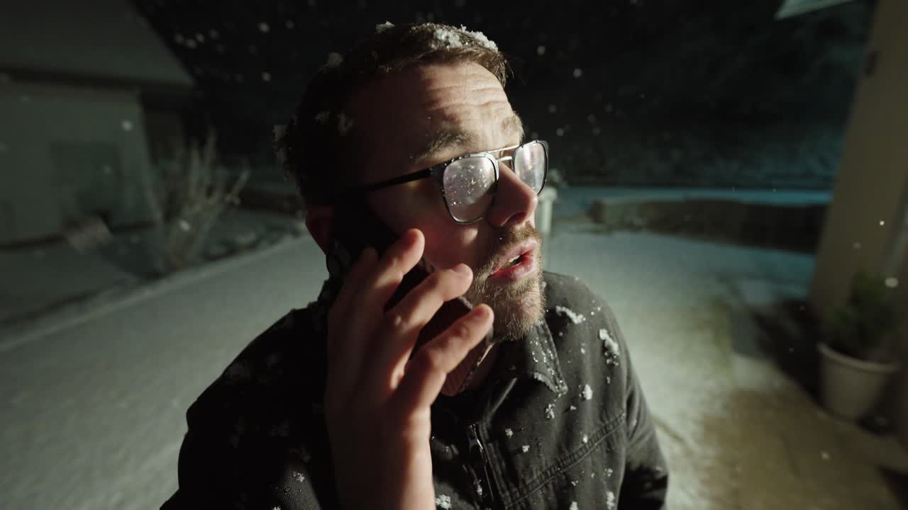 Een man staat 's nachts buiten in hevige sneeuwval en spreekt dringend op zijn telefoon. Concept van blootstelling aan kou, nood, noodcommunicatie en het zoeken van hulp in barre winterse omstandigheden