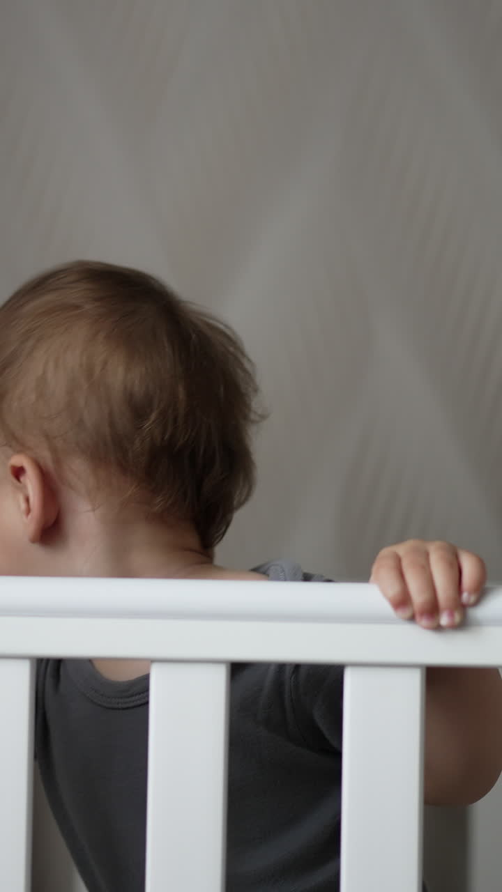 Little boy standing up and sitting down in the cot. Adorable Caucasian child being active after waking up. Vertical video