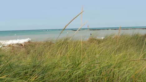 Cerca-De-La-Hierba-De-La-Playa-Que-Sopla-Suavemente-En-El-Viento---Con-Agua,-Surfista-Y-Faro-En-El-Fondo