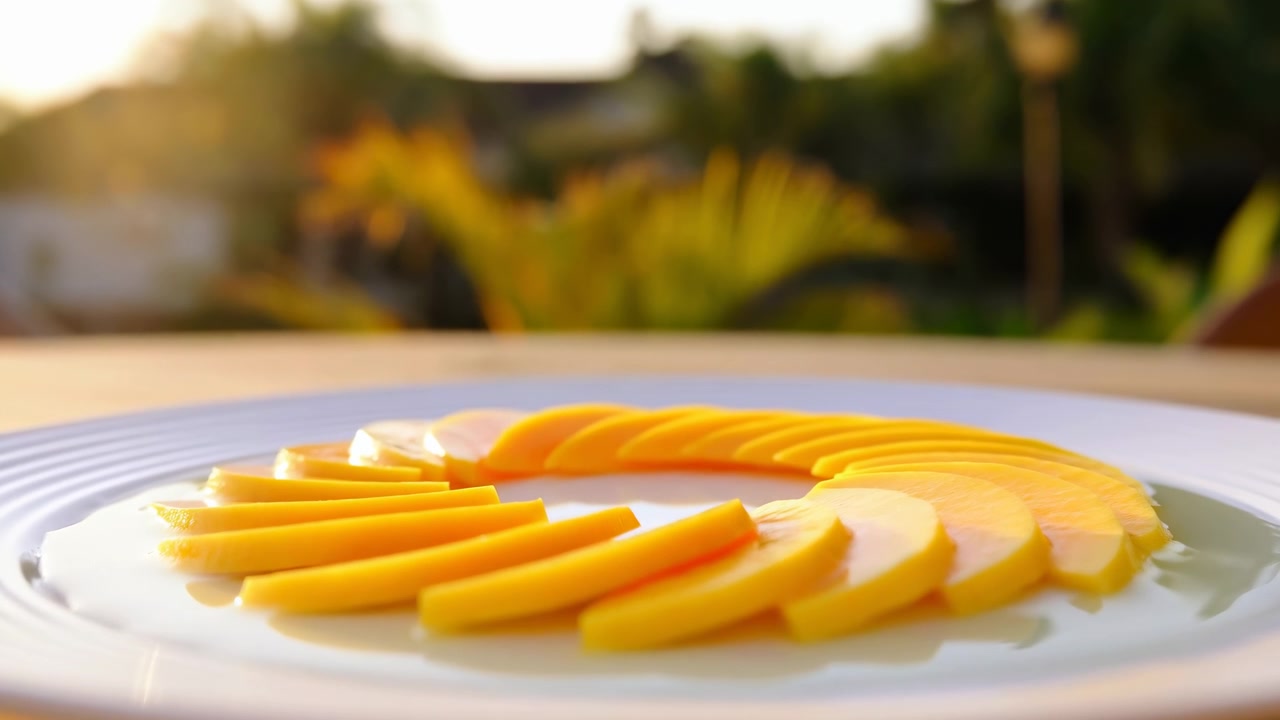 Vista ravvicinata di sottili fette di mango giallo disposte a ventaglio concentrico su un piatto di ceramica bianco scanalato, su un tavolo di legno, con luce solare calda, sfocatura morbida, un'atmosfera tropicale fresca e una presentazione elegante del cibo