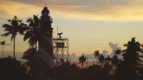 Enfoque-En-Rack-A-Un-Hermoso-Y-Antiguo-Faro-Al-Atardecer-En-Kerala-Con-Cocoteros,-Pájaros-Volando-Y-Una-Torre-De-Radar-Girando-Al-Lado