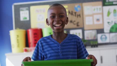 Colegial-Afroamericano-Sonriendo,-Sosteniendo-Una-Papelera-De-Reciclaje,-De-Pie-En-El-Aula