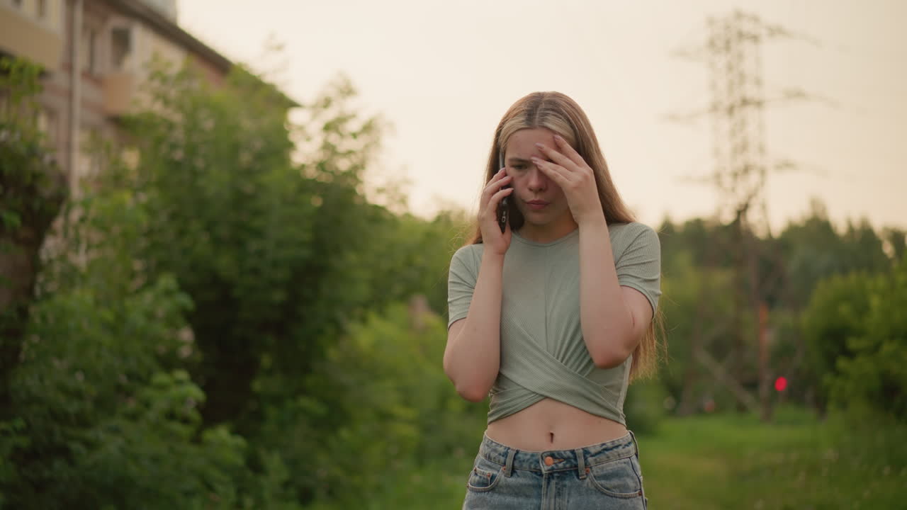 전화 통화를 하는 동안 피곤해 보이는 젊은 여성, 그녀의 왼손은 좌절으로 그녀의 이마를 고, 흐릿한 초록색으로 둘러싸여, 희미한 녹색 빛과 배경에 건물