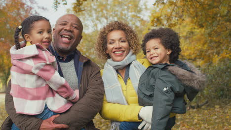 Portrait-Of-Smiling-Grandparents-Carrying-Grandchildren-On-Walk-Along-Track-In-Autumn-Countryside