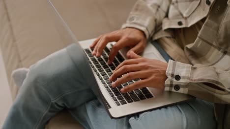 close up of a young man with black skin color in a beige plaid shirt and blue jeans works on a gray laptop and types on the keyboard while working remotely at home on the sofa