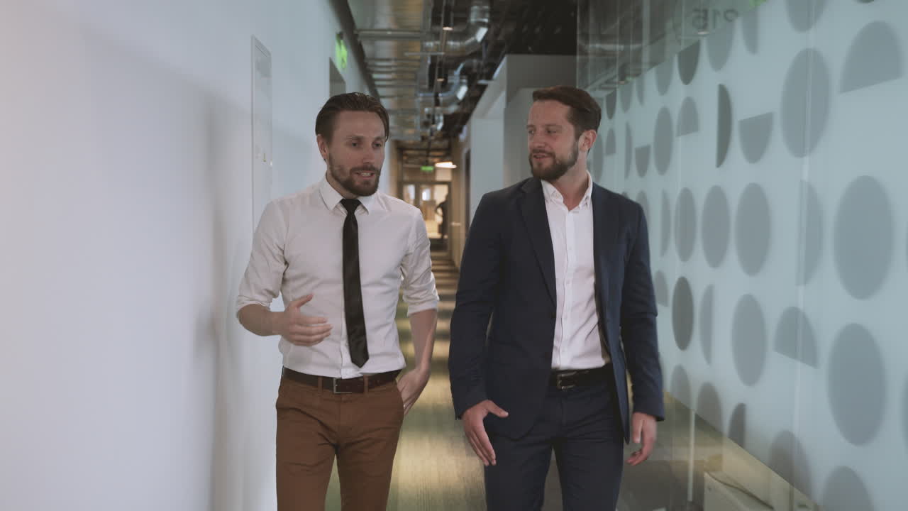 Two young businessmen walking through the corridors of an office building discussing company matters