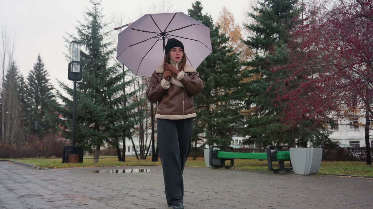 Dreamy girl walking calmly through paved park on cold overcast day while holding closed umbrella, dressed in black knit cap, brown shearling jacket, and black trousers, surrounded by evergreen trees