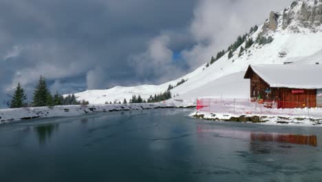 drone panning from the right to the left side of the frame showing a log cabin by a frozen lakeside, at an alpine mountainside in engelberg, switzerland