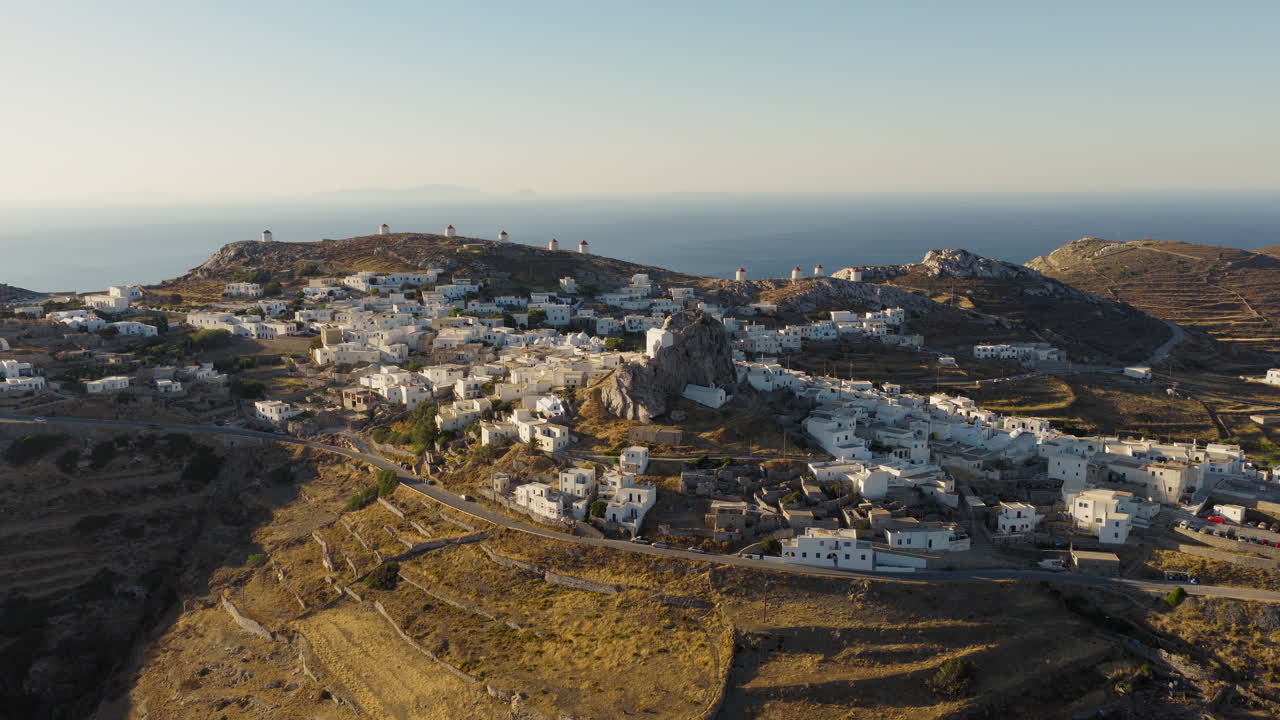 Slow drone reveal of Chora, Amorgos island during sunrise. Soft golden light unveils the whitewashed village with the castle and windmills on the horizon