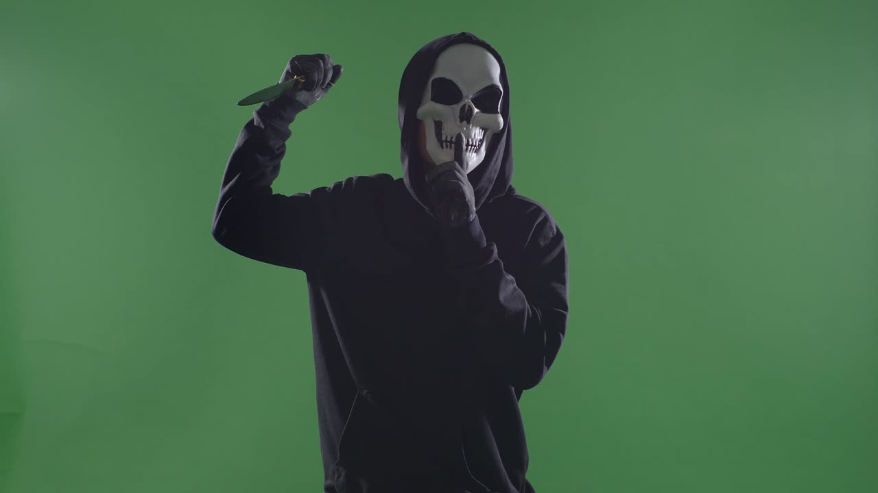 hombre aterrador en la sudadera con capucha con máscara de halloween gestando silencio mientras sostiene un cuchillo en el fondo de la pantalla verde