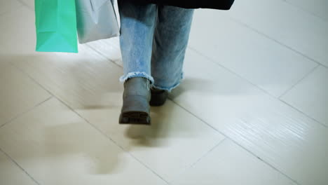 leg view of individual in jeans trousers and boots carrying shopping bag in left hand while walking in mall, shadow cast on tile floor