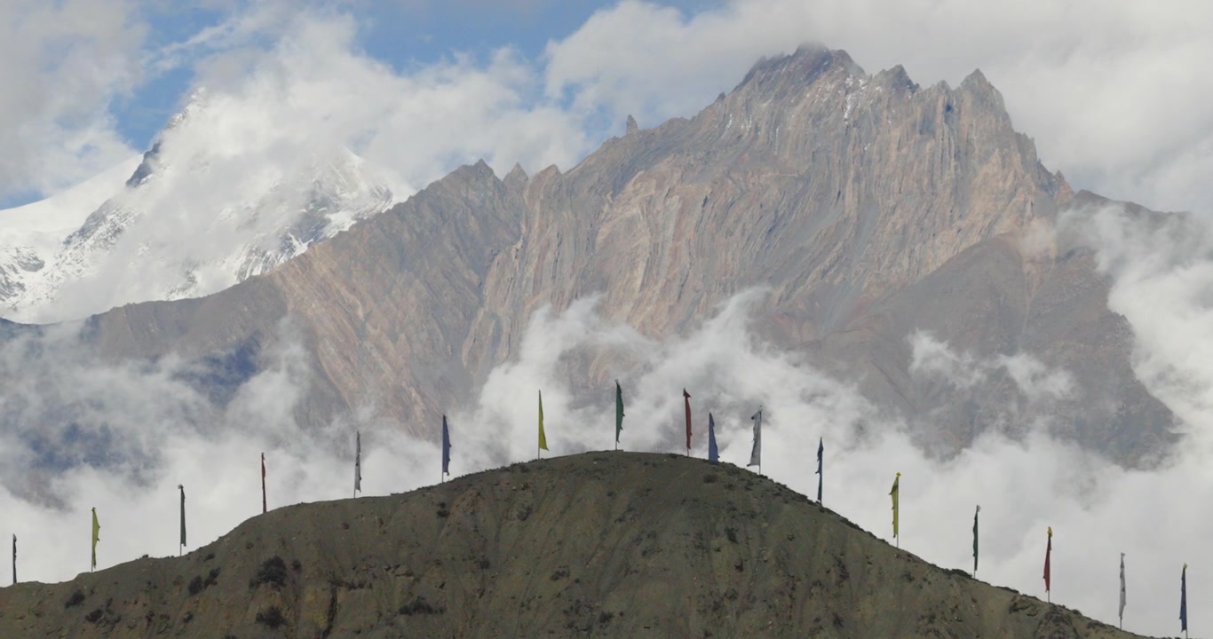 Una ripresa aerea con zoom indietro inizia con un focus sulle montagne himalayane e nuvole basse, muovendosi all'indietro per rivelare una cresta sabbiosa con bandiere di preghiera colorate nel Mustang, Nepal.