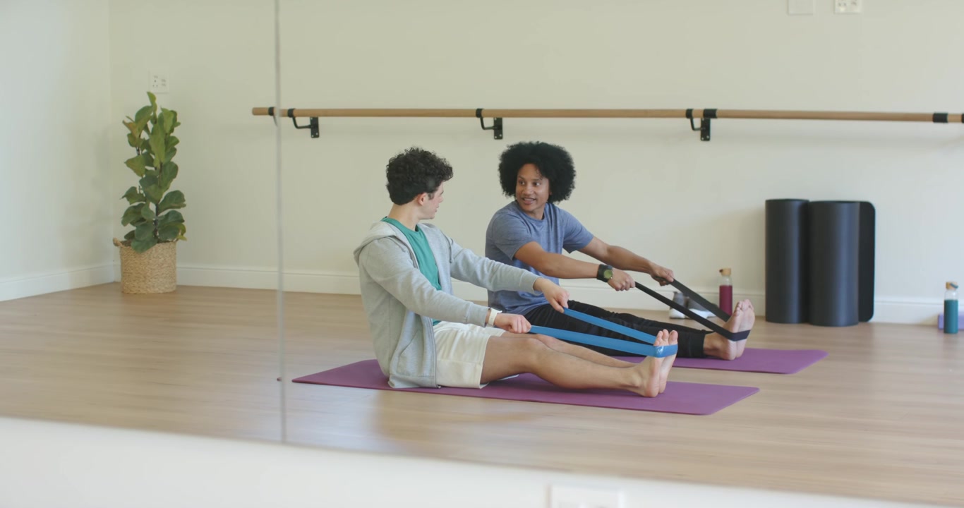 African American male trainer cueing male student pulling bands on mats for seated row by mirror