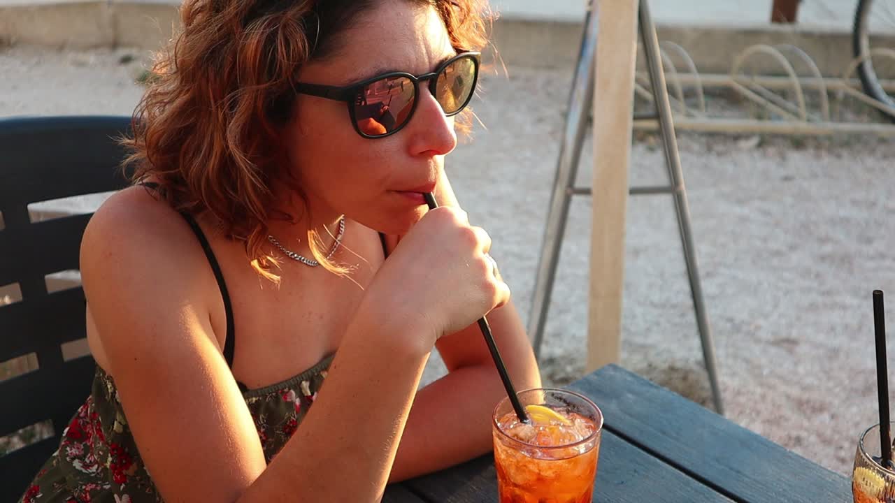 Ragazza con gli occhiali da sole si gode una bevanda colorata al tramonto ventilato.