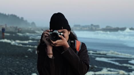 a gorgeous brazilian young woman taking a picture with a vintage film camera at ruby beach in forks, washington during a vivid colorful sunset on a warm summer evening