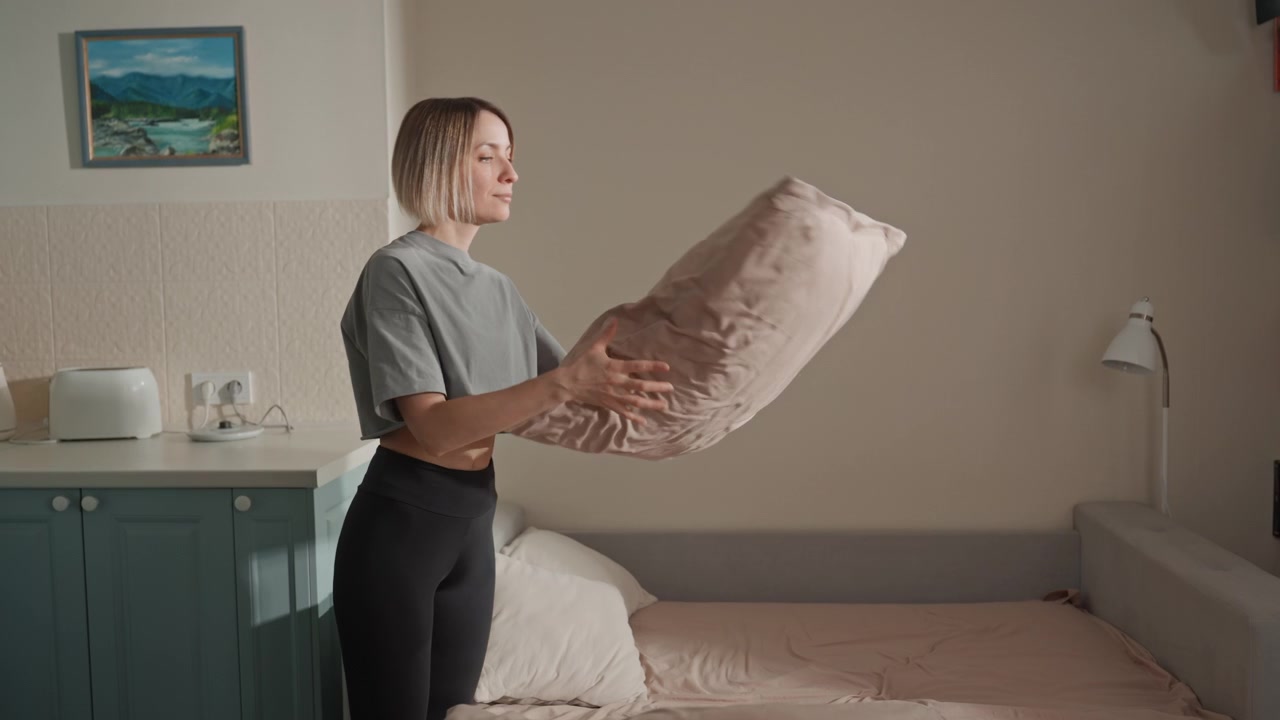 Donna bianca che sistema il cuscino in camera da letto, luce naturale soffusa, biancheria da letto color pastello, sorriso sereno, movimenti ritmici che suggeriscono un umore da amante della musica, top grigio casual, lampada da comodino, quadro paesaggistico incorniciato.