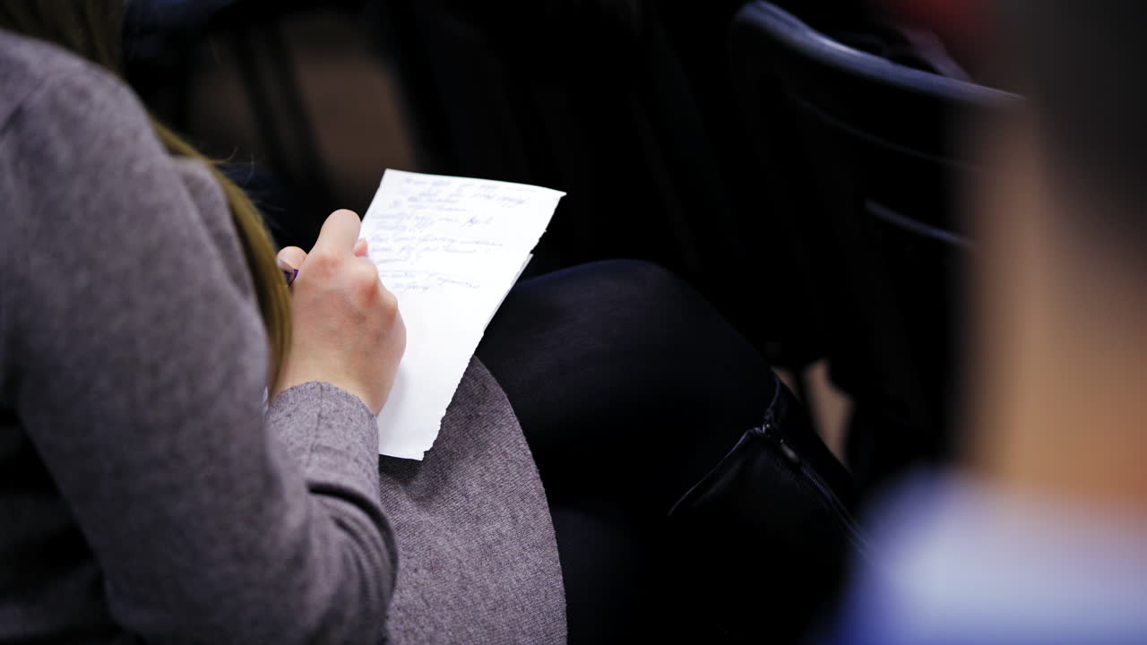 Young female is writing on a sheet of paper. Woman is sitting in a conference hall and notes some information. Close-up.
