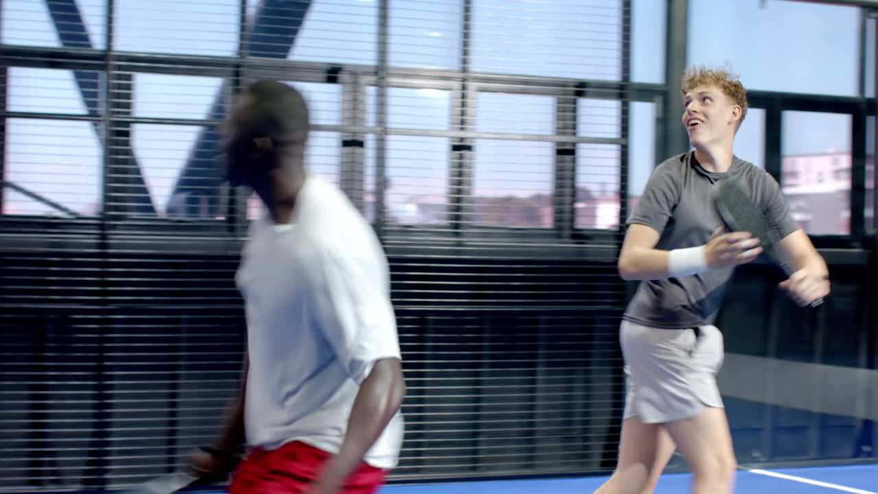 Two diverse men walking on padel on indoor court holding rackets, ready for match
