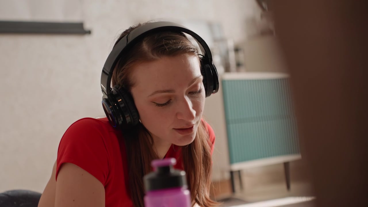 Giovane donna in sessione di allenamento a casa con cuffie, maglietta rossa, borraccia viola visibile, in un soggiorno interno con luce naturale soffusa, che segue l'audio di un trainer online, con respirazione concentrata e movimenti delicati