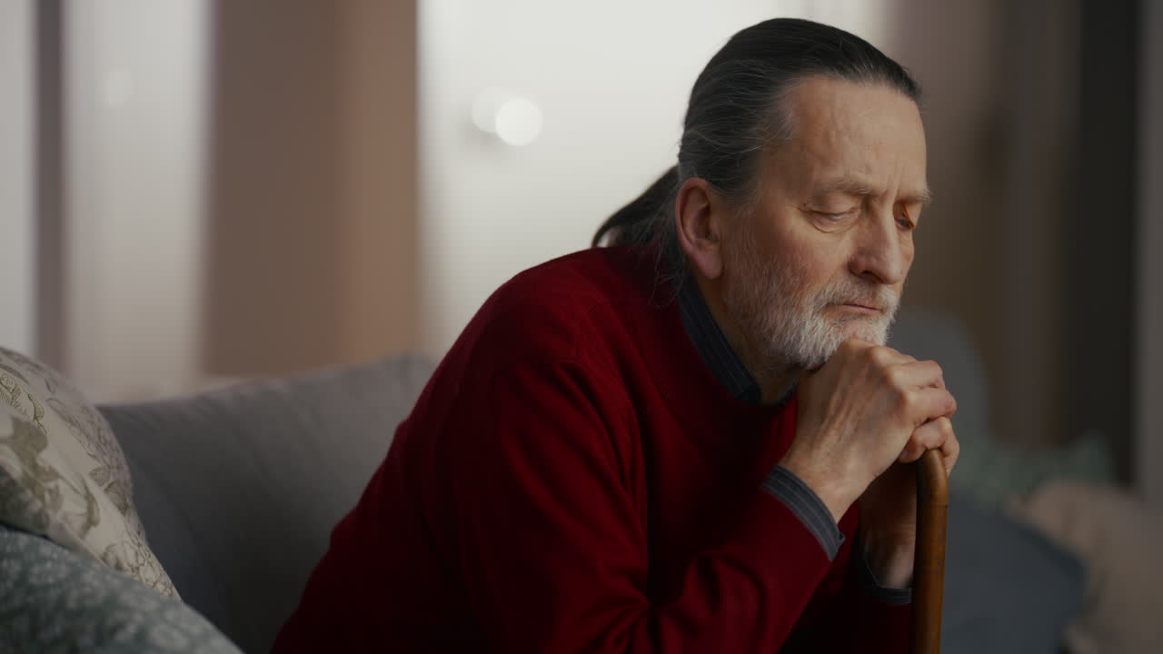 Thoughtful Elderly Man with Walking Stick Sitting on Couch