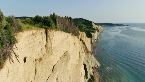 Cliffside-forest-on-Corfu-Island-overlooking-the-Ionian-Sea,-sunny-day,-aerial-view