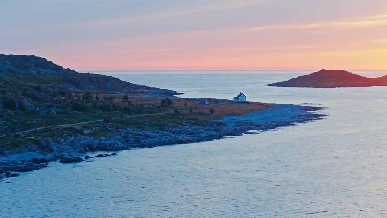 Sandland, Loppa Kommune, Finnmark Fylke, Norge - Et ensomt hus ligger på en stenet kystlinje med udsigt over det rolige hav under en blød pink solnedgangshimmel - Orbit droneoptagelse.