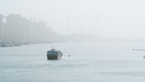 Vögel-Fliegen-über-Dubai-Creek-Mit-Fischerboot-Und-Stadt,-Die-Von-Dichtem-Nebel-In-Deira,-Dubai,-Vereinigte-Arabische-Emirate-Verdeckt-Werden