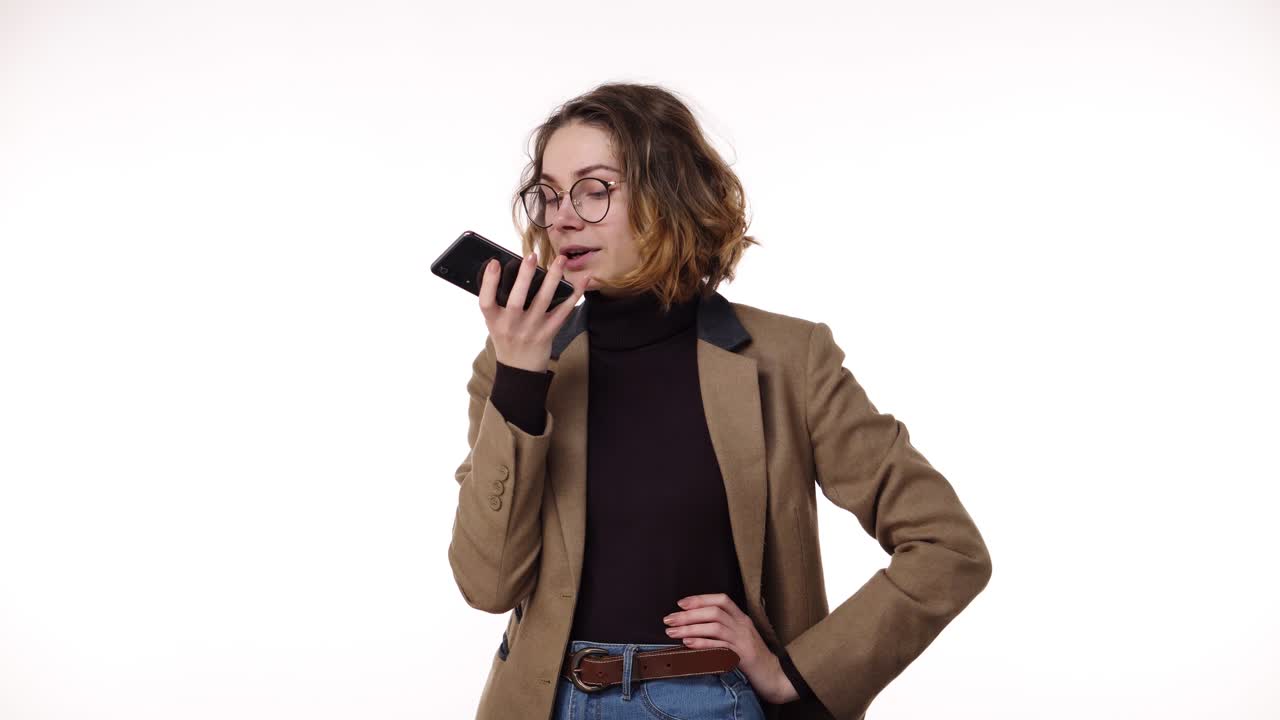 Beautiful brunette woman in stylish casual clothes records a voice message on her mobile phone on white background in studio. Talking emotionally to her friend