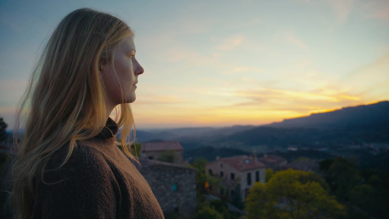 A close side profile of a woman catches the last soft sunset light above Siurana, Spain, while blurred rooftops, distant hills, and pastel evening color create a calm and intimate atmosphere.