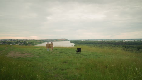 an artist in a black hat and plaid shirt stands close to a woman in a hat, both focused on a drawing board set up in a lush grassy field. a lake and a lone chair add depth to the tranquil scene