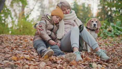 woman gently places hand around sad looking daughter sitting close in quiet autumn forest while dog calmly sits beside them surrounded by fallen leaves and warm earth tones