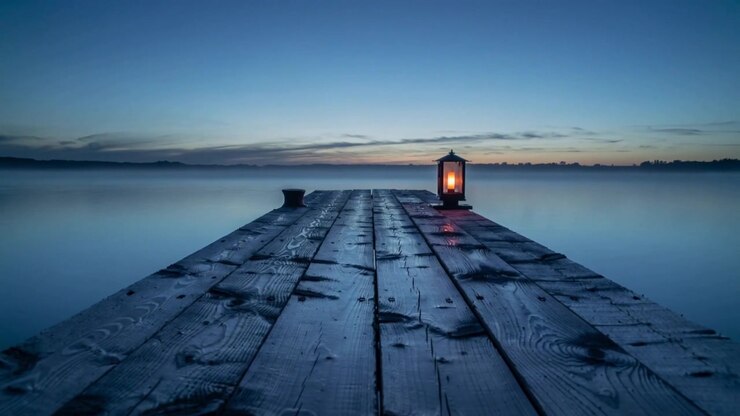 Panning camera capturing wet wooden pier reaching calm lake at dusk, highlighting lantern glow
