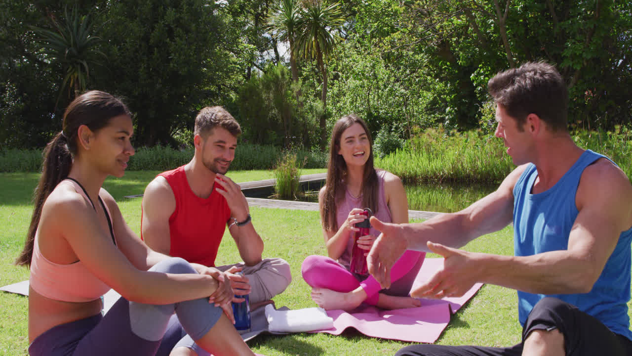 instructor de yoga masculino hablando con un grupo diverso feliz sentado en esteras en un parque soleado