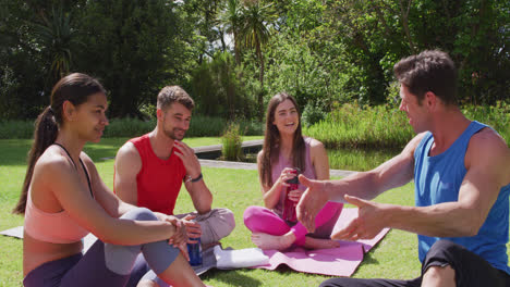 male yoga instructor talking with happy diverse group sitting on mats in sunny park