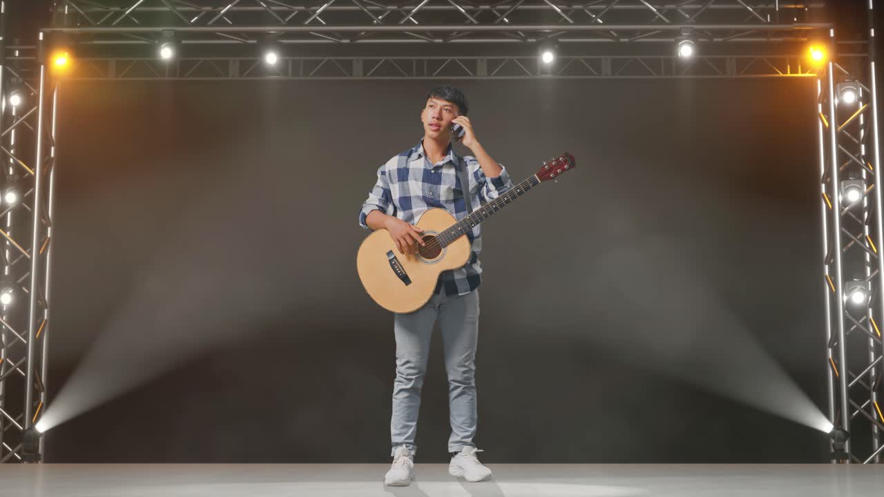 cuerpo lleno de adolescente asiático con guitarra hablando en teléfono inteligente mientras está de pie en el escenario