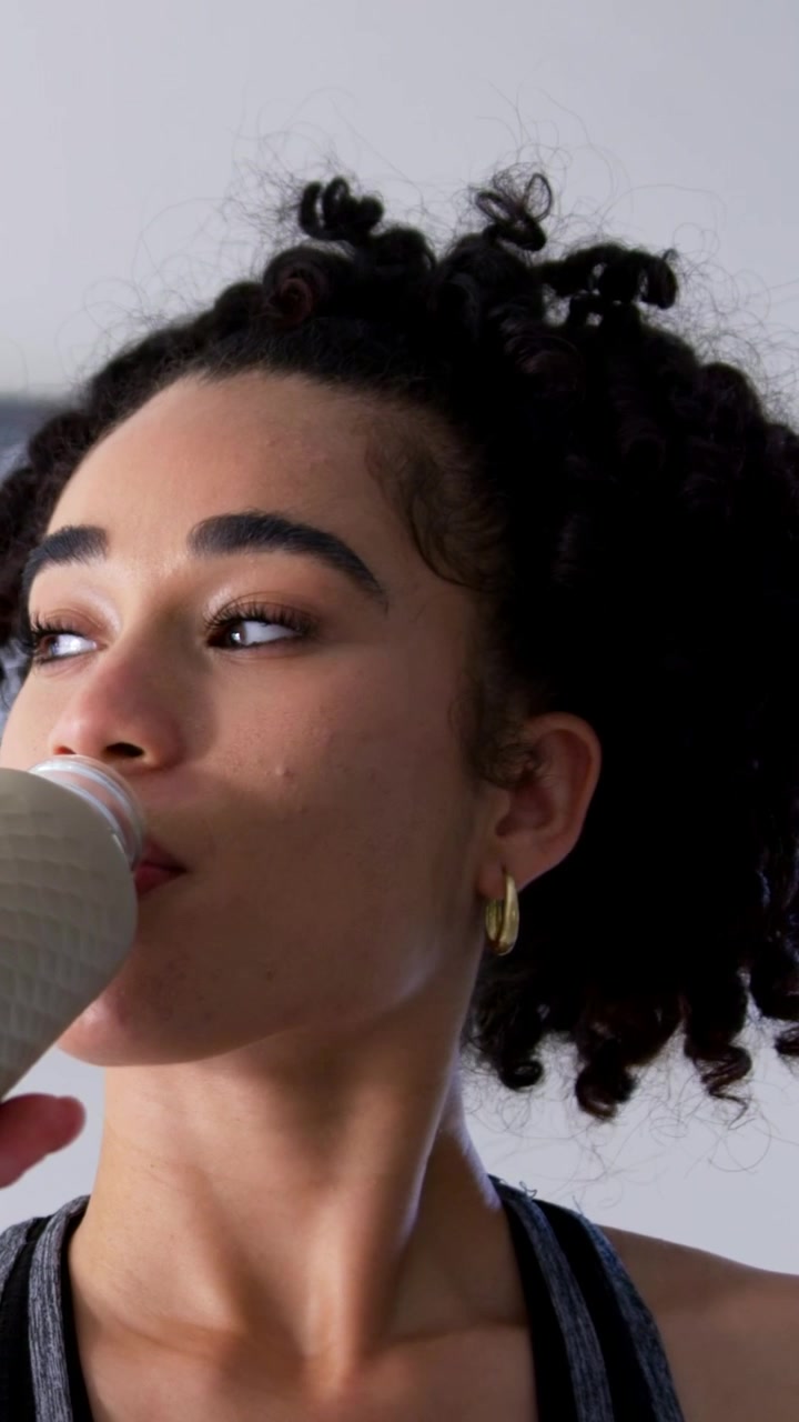 Vertical video: Drinking woman after workout, lifting textured bottle, sipping in room, in tank top