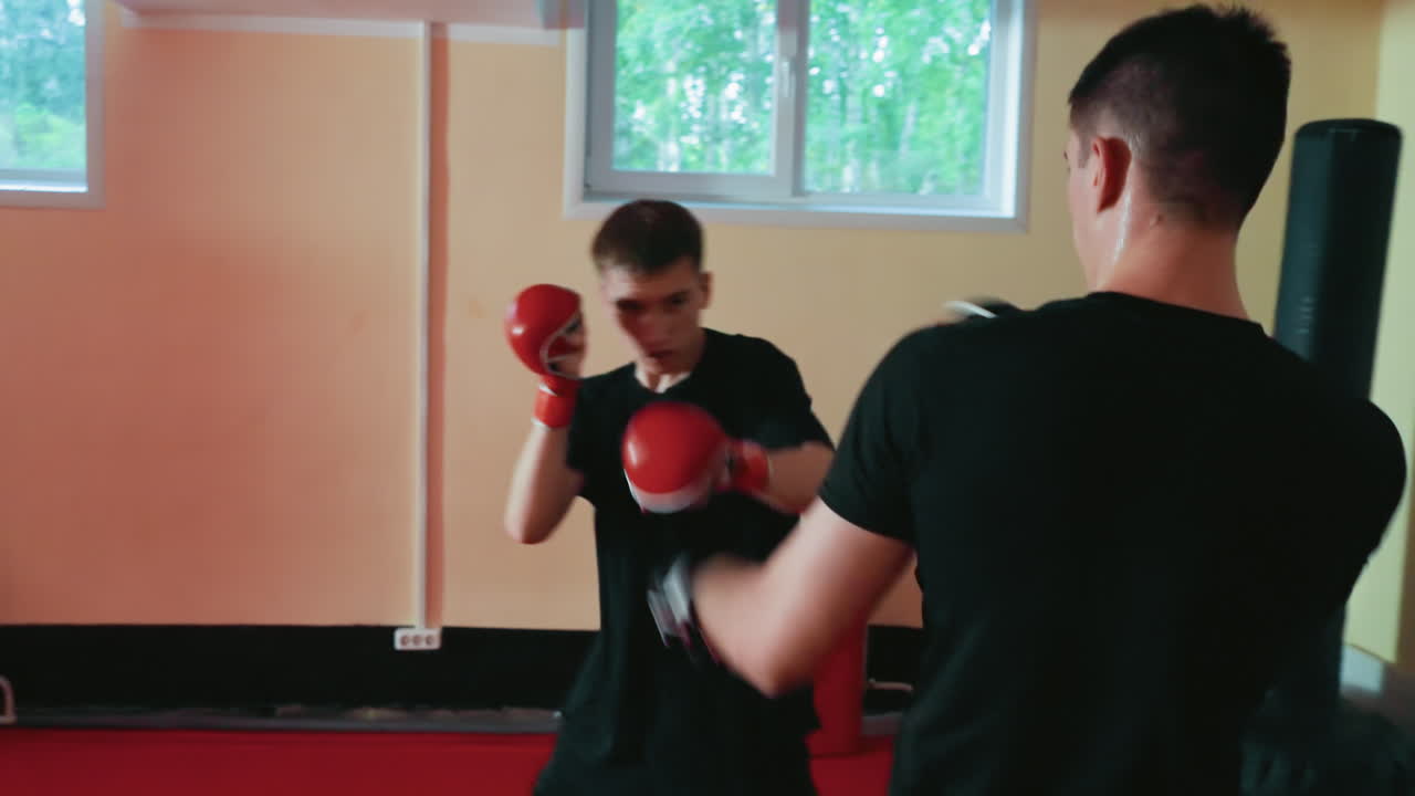 Fighters engaged in sparring session, young man in red gloves throwing punch toward opponent in black gloves inside martial arts gym, showcasing technique, intensity, focus, strength, endurance