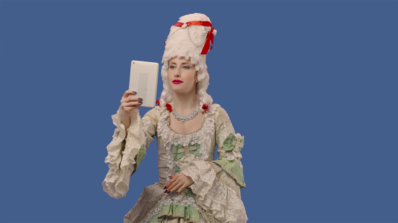색 빈티지 드레스와 위그를 입은 궁정 여성의 초상화, 디지털 태블릿을 사용하여 비디오 채팅을 위해 대화하는 젊은 여성, 블루 스크린 배경으로 스튜디오에서 포즈를 취하는 젊은 여성.