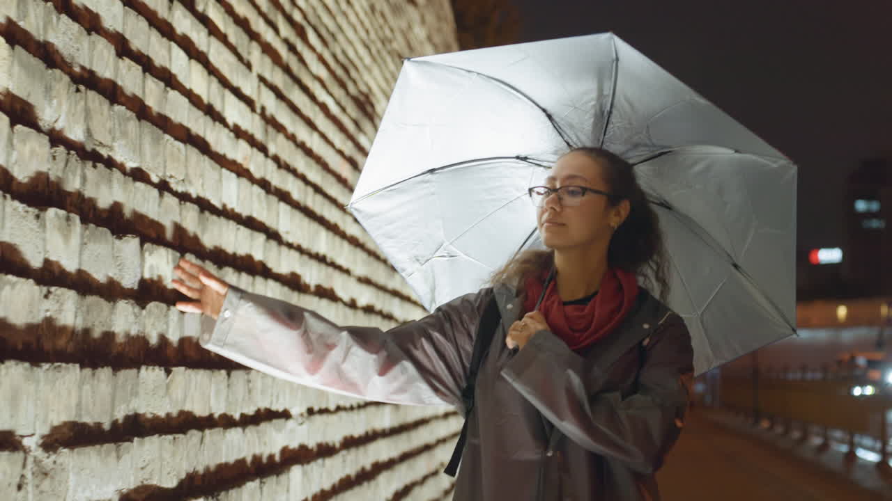 Donna asiatica con ombrello che tocca una parete illuminata, indossando un impermeabile e una sciarpa rossa, la delicata punta delle dita traccia il cemento testurizzato, scena di strada notturna cinematografica e malinconica, bagliore di un'installazione luminosa, silenziosa.