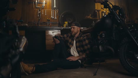 Happy-guy-mechanic-with-a-beard-is-browsing-social-networks-during-his-relaxation-in-a-motorcycle-workshop