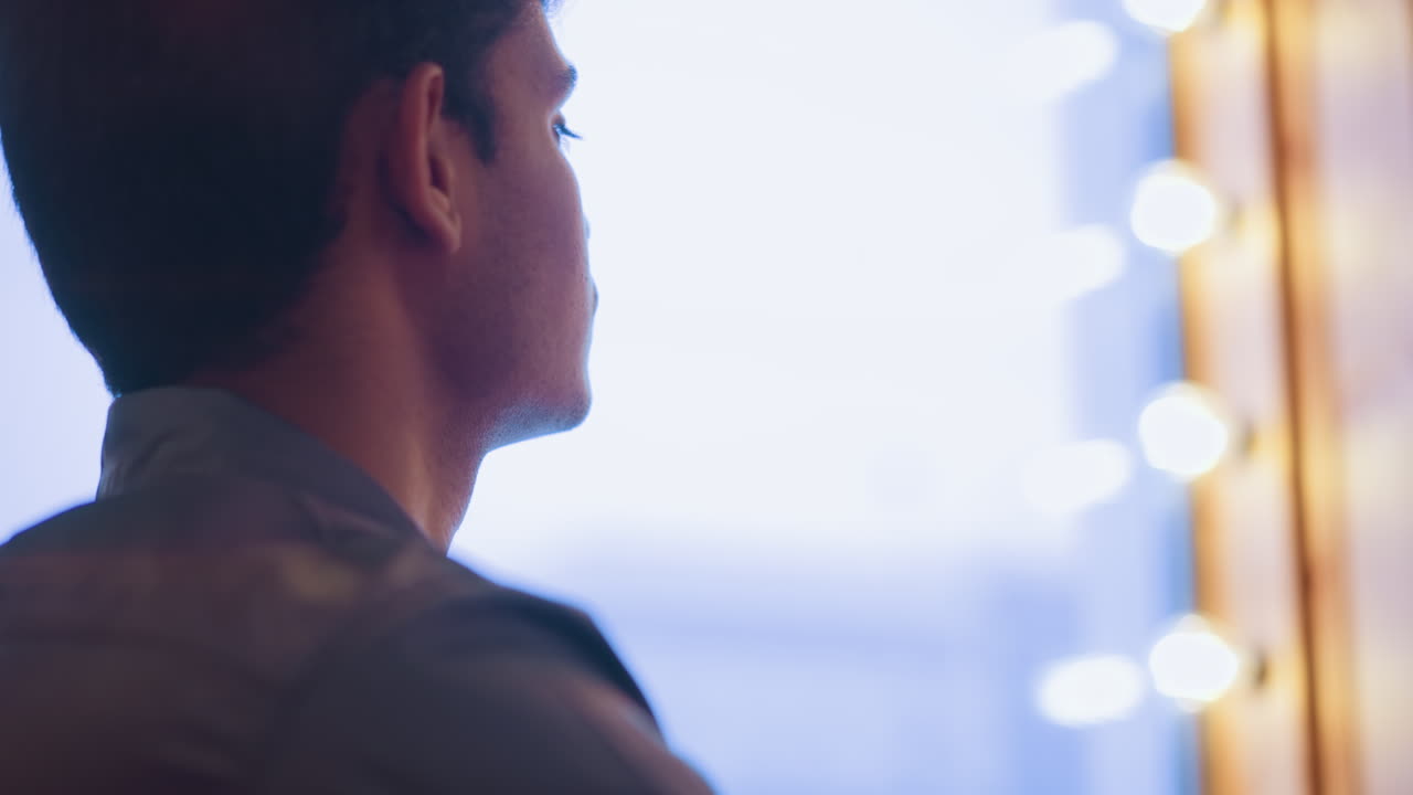 Close-up of young man looking thoughtful while sitting indoors near brick wall, illuminated by soft ambient lighting, conveying introspection, solitude, and quiet emotional depth in urban setting