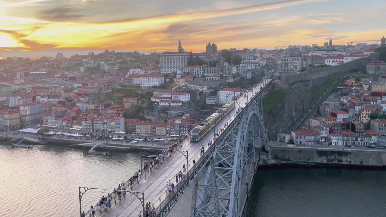 Aerial footage shows metro train moving across Dom Luís I Bridge at sunset, with vibrant rooftops and historic buildings of Porto in background beside calm Douro River under colorful evening sky