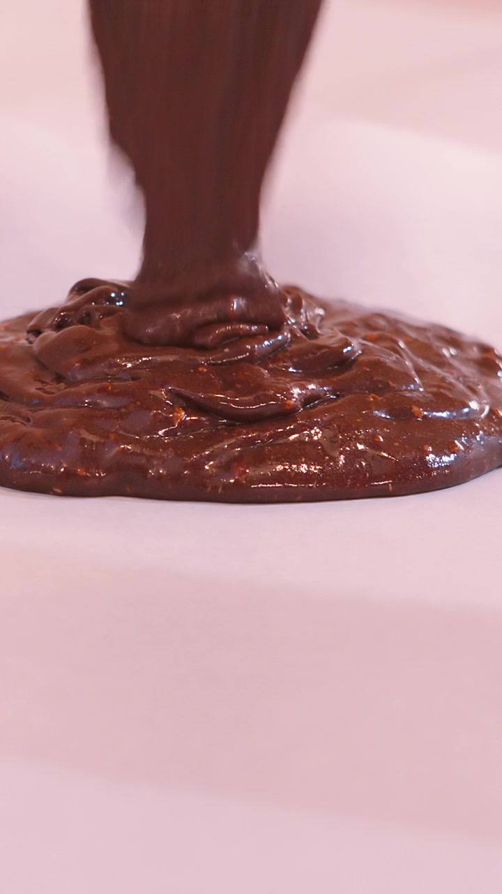 Video verticale che versa densa pastella per brownie al cioccolato su carta da forno in una teglia. La pastella al fudge forma nastri e si ripiega su se stessa mentre scende, un'immagine appetitosa di un dessert fatto in casa