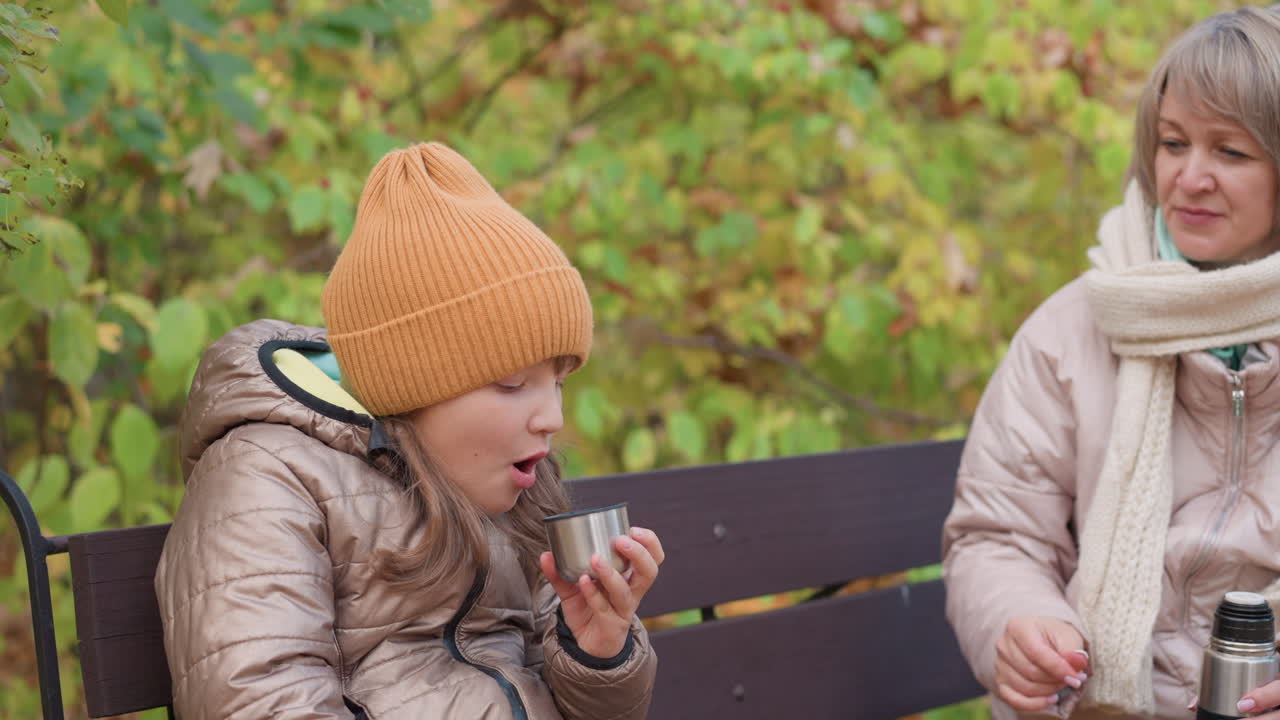 Mother seated on park bench hands warm drink in metal cup to daughter blowing on it gently to cool as both wear cozy jackets surrounded by golden autumn leaves in serene outdoor setting
