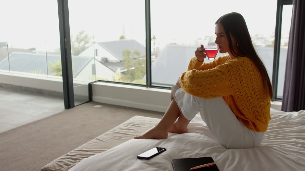 Relaxing at home, woman drinking tea while sitting on bed with smartphone